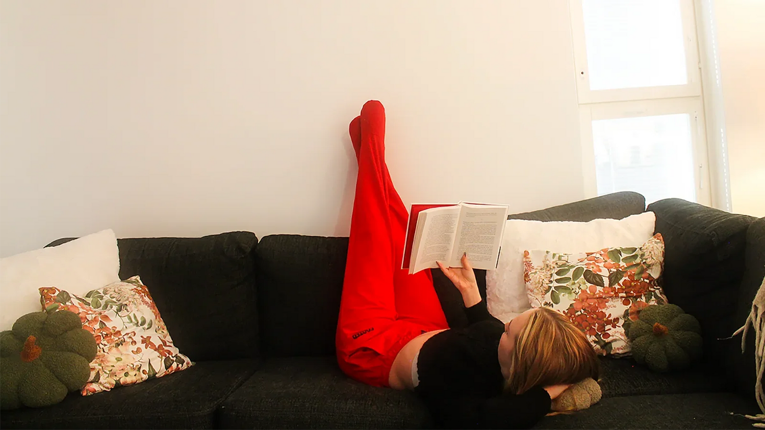 Person wearing red FOOTED pants lying on a couch with legs up, reading a book — a cozy moment of comfort and calm.