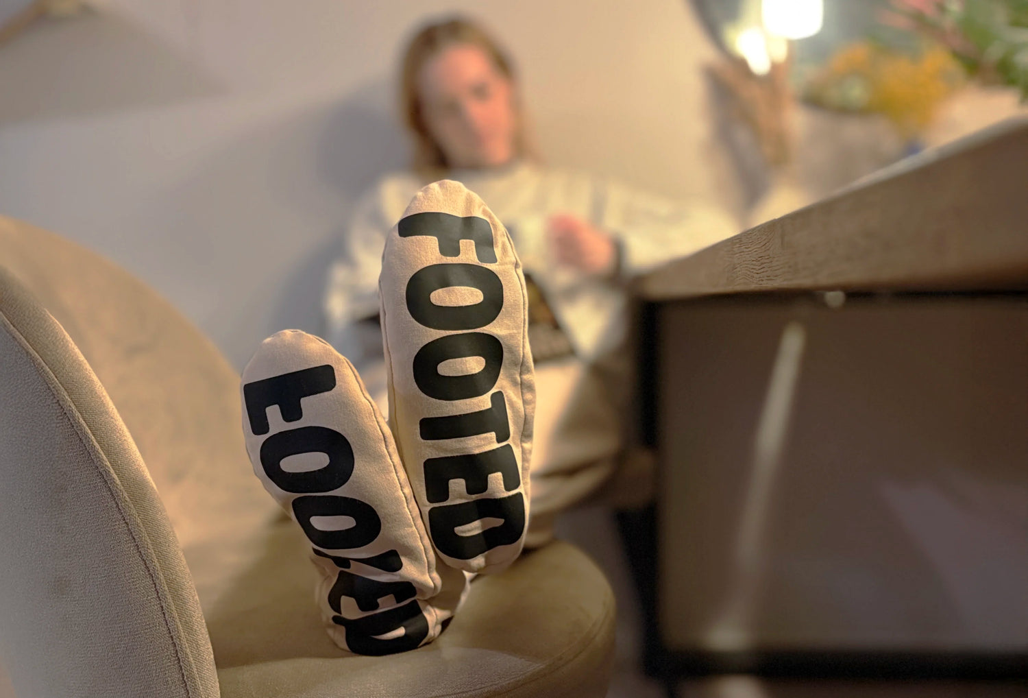Person relaxing indoors wearing beige FOOTED pants, with focus on the soles showing the logo.