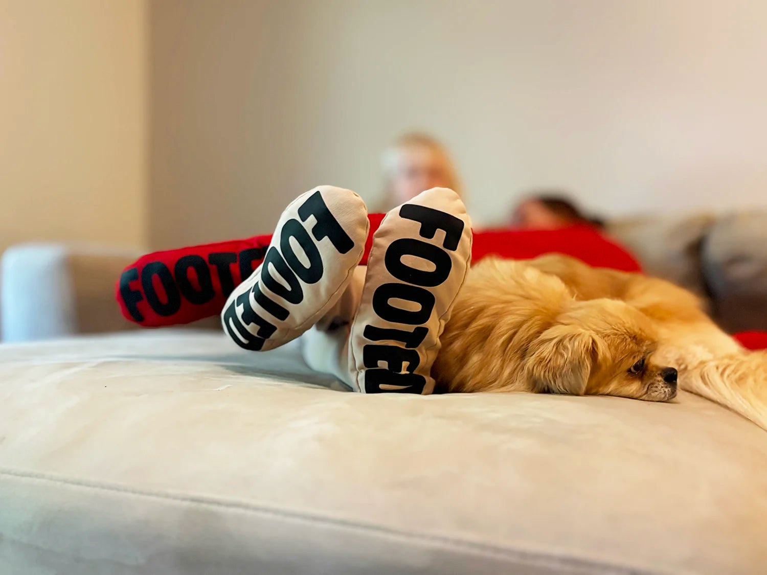 Person relaxing on a couch wearing beige and red FOOTED pants, with a small dog resting beside them.