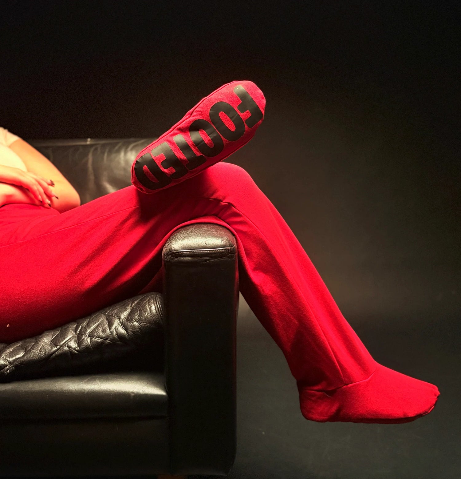 Red FOOTED pants with footed design, worn while sitting on a black leather chair against a dark background.